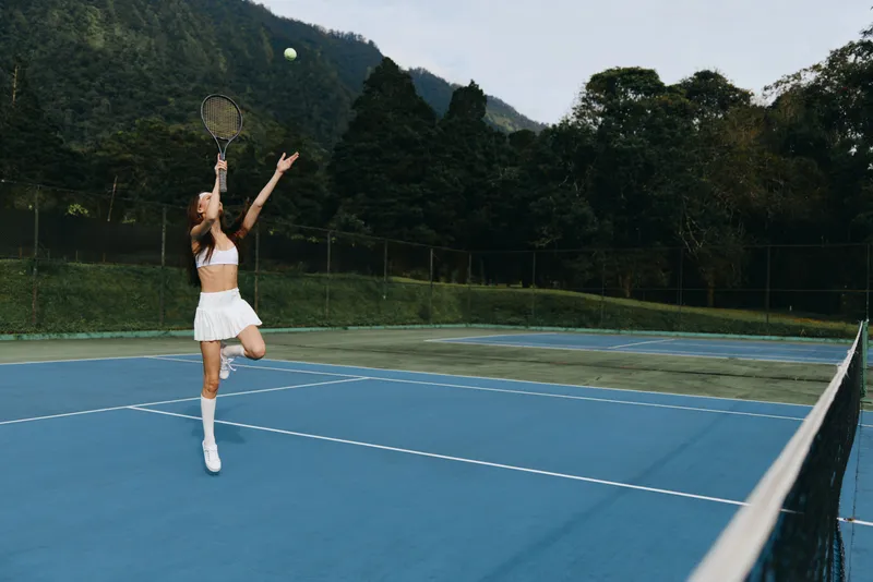 Young female tennis player serving on a blue court, showcasing athleticism and determination in a scenic outdoor setting