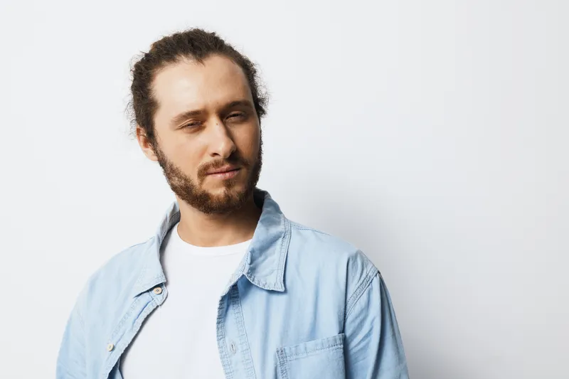 Confident young man with curly hair wearing a light blue shirt, expressing skepticism against a plain white background, ideal for lifestyle projects