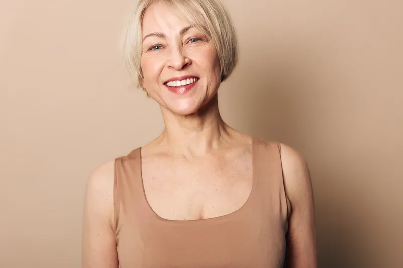 Happy mature woman with short blonde hair smiles warmly wearing beige tank top. Studio portrait on neutral background. Concept of aging gracefully, confidence, and natural beauty.