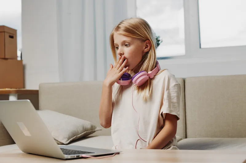 Blonde girl with pink headphones sits on sofa looking surprised at laptop screen in bright living room. Concept for remote learning, online discovery, and childhood curiosity.