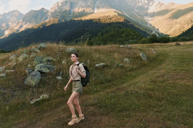 Young woman hiking outdoors in mountain landscape, casual outfit, carrying backpack, enjoying nature, sunny day, green grass and rocky terrain