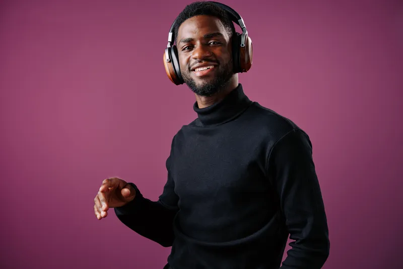 Young African man smiling with headphones, wearing a black turtleneck against a purple background, embodying joy and passion for music