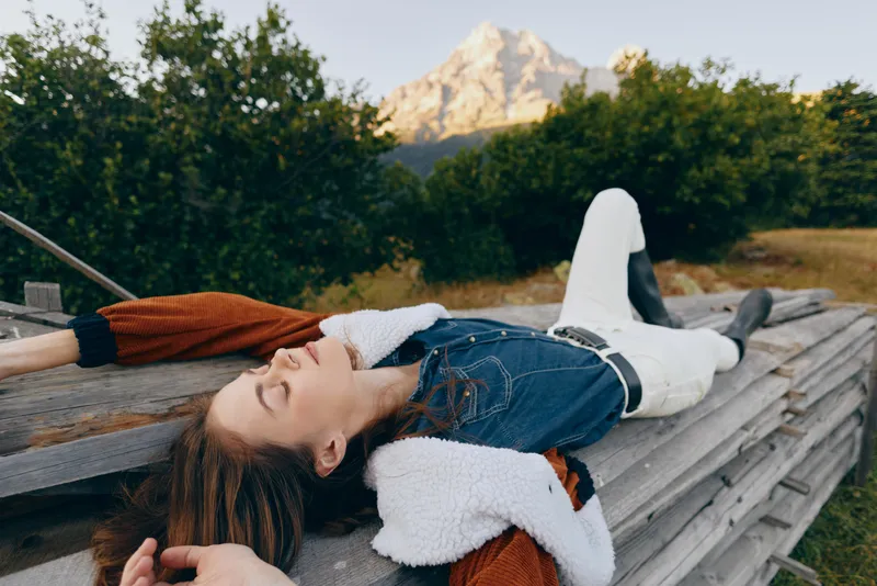 woman relaxation nature mountain meadow denim, young woman lying on wooden platform in countryside with eyes closed, casual denim jacket and white pants, serene outdoor portrait with distant peak