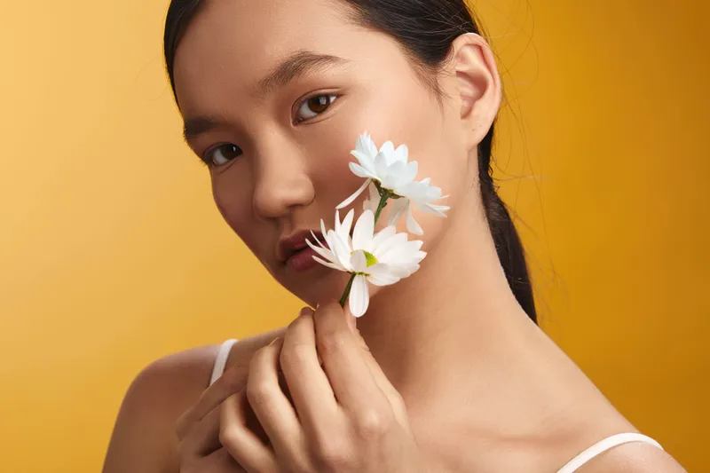 Asian woman holds white flowers near her face against a warm yellow backdrop, creating a serene portrait that emphasizes natural beauty and a calm, graceful mood.
