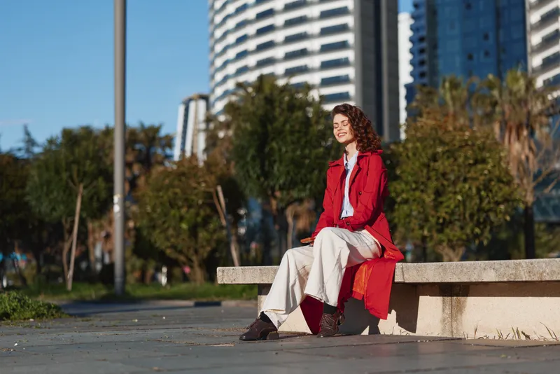 One person, young woman with curly hair sits on a concrete bench in an urban park, wearing a red coat and white pants, enjoying a sunny day and calm surroundings.