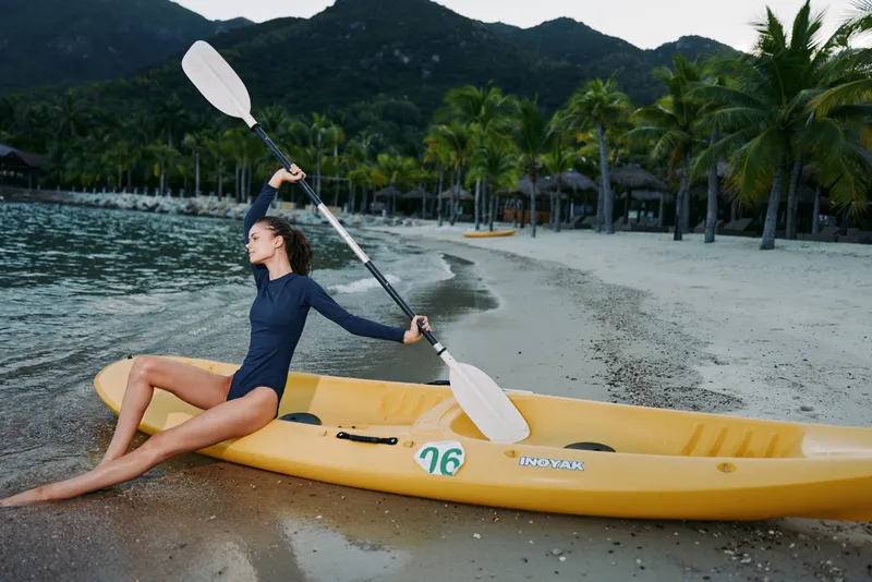 A kayaker sits on a bright yellow kayak at the shoreline, raising a paddle as small waves roll in and distant mountains frame the tropical scene.