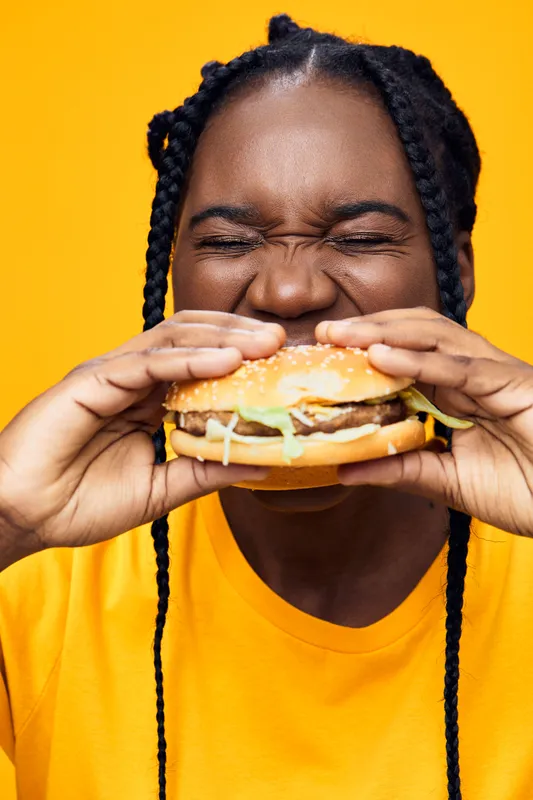 Happy woman enjoying a delicious hamburger, wearing a bright yellow shirt against a vibrant yellow background, showcasing joy and a love for food