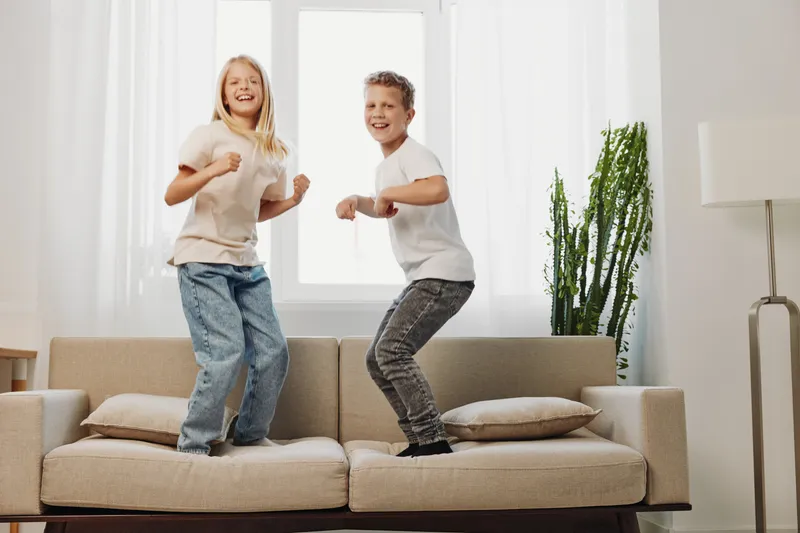 Happy children jumping on sofa in bright living room, smiling and having fun together. Concept of childhood joy, family time, energy, and playful indoor activity for kids.