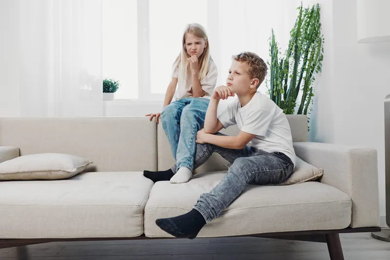 Two children sit on a beige couch in a bright living room, showing thoughtful and pensive expressions. Concept of sibling emotions and family dynamics.
