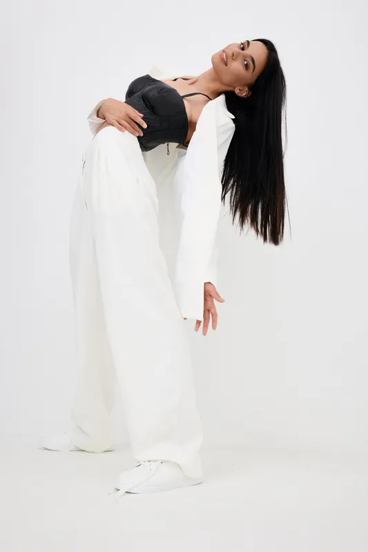Young woman wearing a black corset and white wide-leg pants posing dynamically against a white background, showcasing modern fashion and elegant movement.