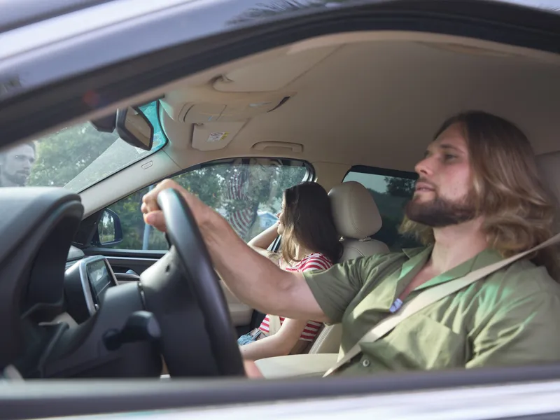 Casual car interior scene featuring a man driving with both hands on the wheel while a passenger sits in the back, soft daylight creating a relaxed mood.