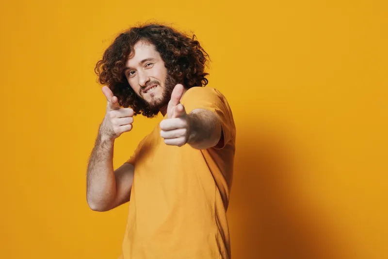 Cheerful young man with curly hair in a yellow t shirt posing playfully against a vibrant yellow background, expressing confidence and joy
