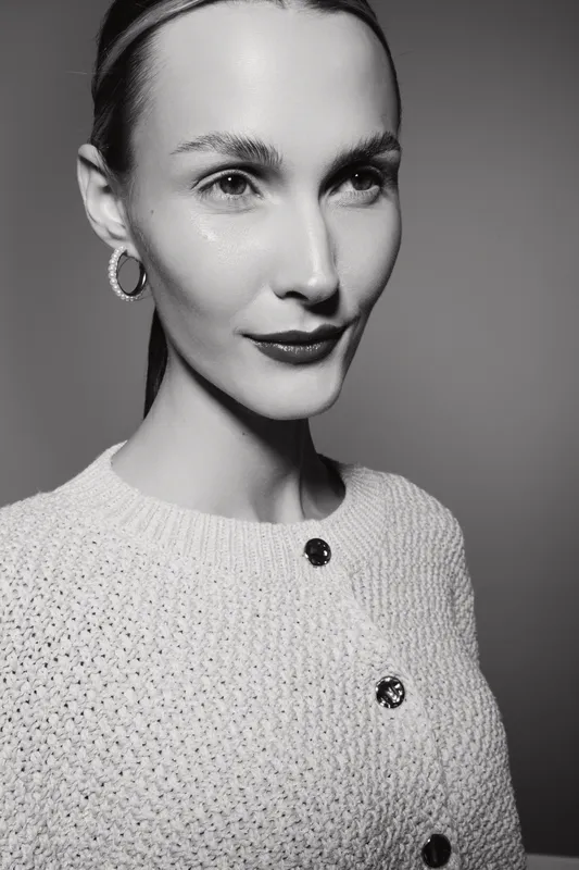 Portrait of a Caucasian woman with fair skin wearing a knitted sweater and hoop earrings. The black and white image highlights her serene expression and smooth complexion.
