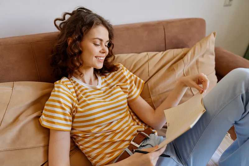 A young woman with curly brown hair wearing a striped shirt relaxes on a cushioned couch, reading a paper in a cozy living room, conveying a quiet everyday home moment of relaxation.