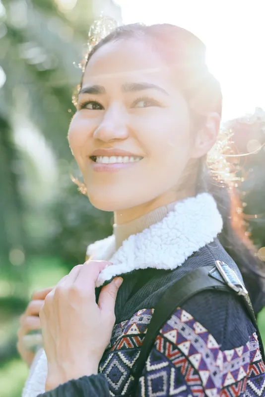 Smiling woman outdoors with towel draped over jacket, sunlit background and greenery, casual fashion, candid moment captured during a day out, fresh air and relaxed vibe suitable for lifestyle,