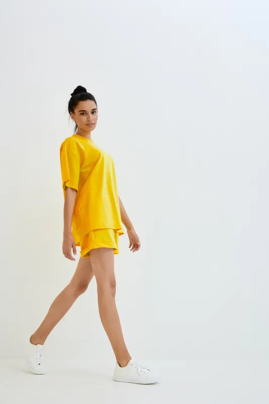 Young woman wearing bright yellow casual outfit and white sneakers walking confidently against minimal white background indoors, modern and fresh style.