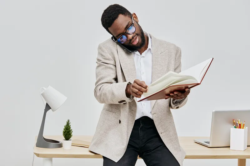 Confident man with glasses reading a book while standing in a modern office space, dressed in smart casual attire, smiling and enjoying leisure activity, creative workspace, bright lighting, professio