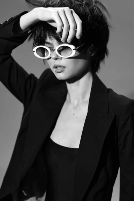 Black and white portrait of a stylish woman wearing round sunglasses and a blazer with short hair and a confident expression.