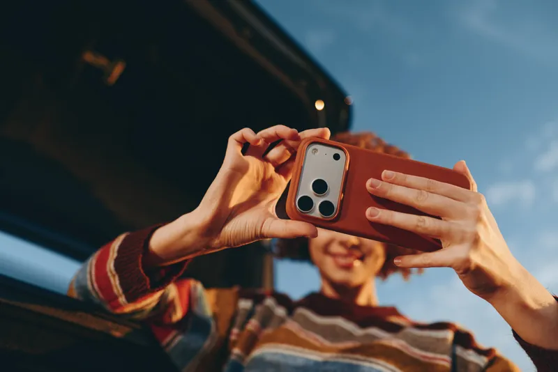 Selfie moment, woman holds smartphone with two hands, friends outdoors near car, smiling and taking picture in road trip lifestyle, sunny sky, casual travel scene