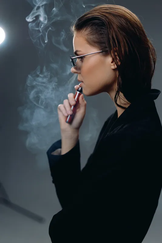 Stylish woman vaping with eyeglasses, exhaling smoke in dark modern background.