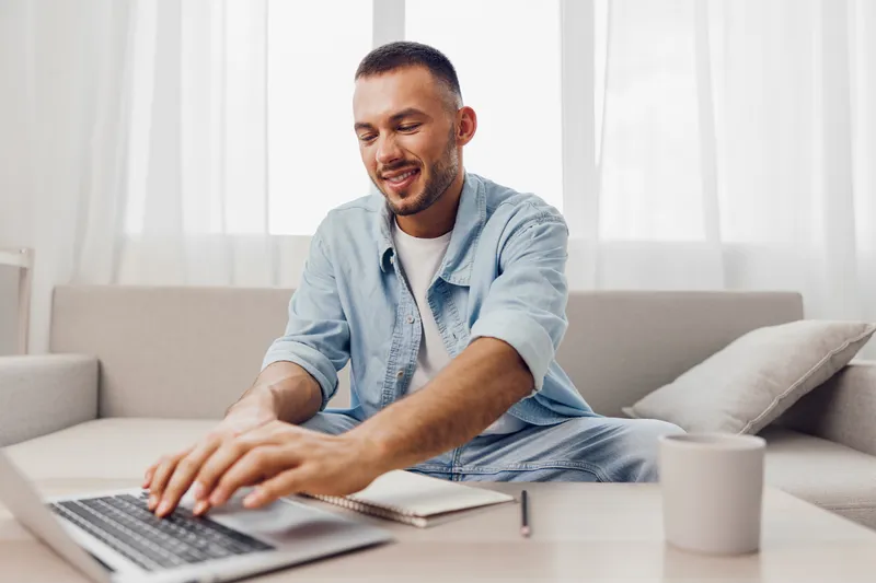 Smiling man focused on his laptop, casually dressed in a denim shirt, enjoying a productive work-from-home environment with natural light and minimalistic decor.