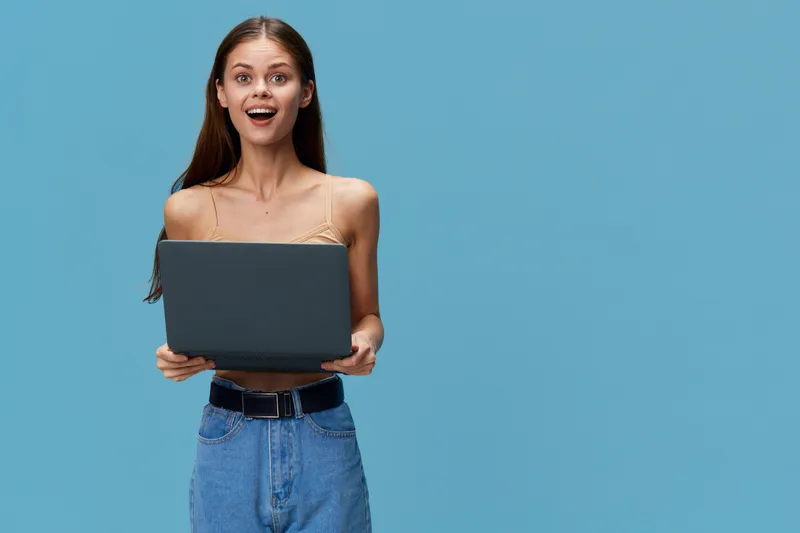 young woman holding laptop with surprised expression, casual clothing and blue background, technology and digital concept in modern lifestyle