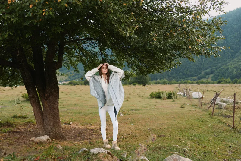 A woman in light clothing stands in a rural field under a large tree, hands on her head, capturing a relaxed outdoor lifestyle moment in nature.