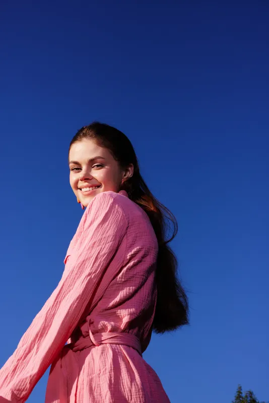 Cheerful young woman in a pink dress against a clear blue sky, exuding happiness and vitality in a vibrant summer setting