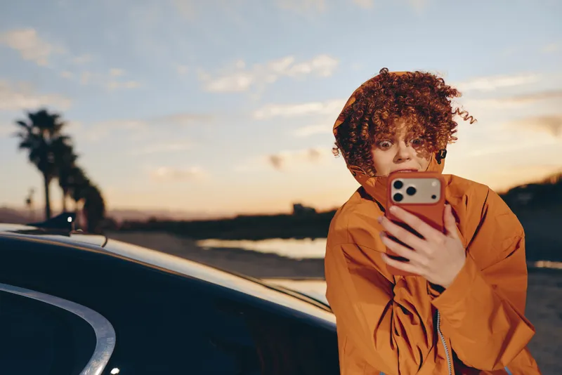 Woman with curly hair takes a selfie with a smartphone near a car outdoors at sunset, warm lifestyle mood, casual travel scene, soft natural light, golden evening sky in background
