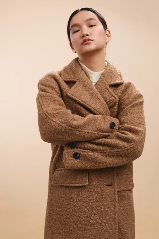 asian woman in a brown wool coat stands with arms crossed in a studio, projecting elegance, confidence, and a modern fashion vibe through a poised portrait, set against a soft warm backdrop and clean