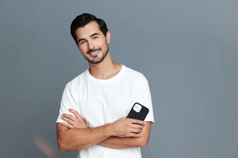 Smiling man with crossed arms holds smartphone while standing against a plain gray background. Casual style and relaxed mood add to the cheerful atmosphere.
