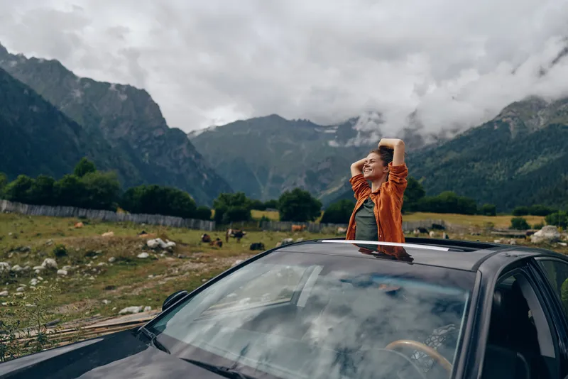 Person standing through the sunroof of a car in a dramatic mountain landscape, celebrating travel outdoors with a joyful expression, capturing adventure and freedom during a roadside vacation.