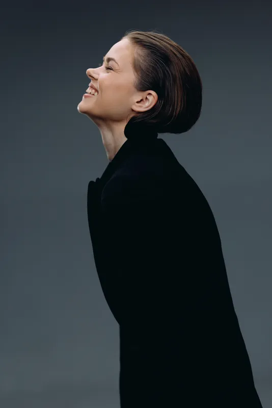Smiling woman with short hair wearing black clothes, showing happiness and confidence against a dark gray background.