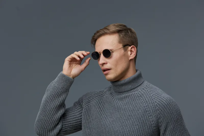 Modern young man wearing round sunglasses and gray knitted turtleneck look serious and stylish against a plain gray background studio portrait. Fashion and style concept
