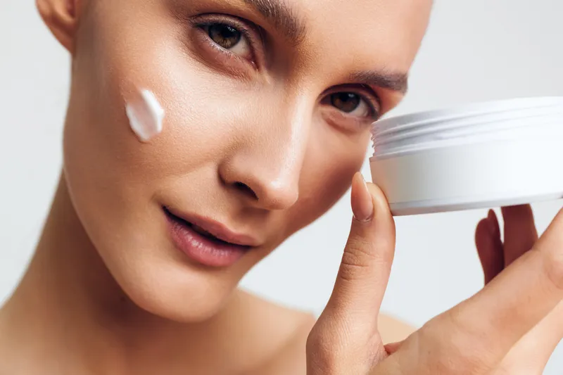 Close-up of a woman applying white face cream on her cheek. Clear skin and natural makeup emphasize skincare and beauty routine with soft lighting on a white background.