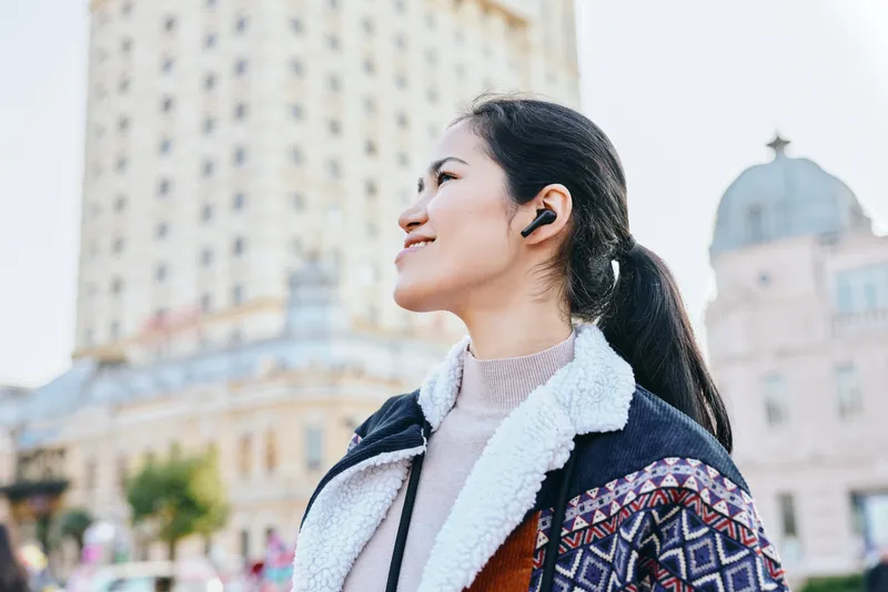 woman headphones city outdoor jacket listening urban fashion lifestyle street scenery skyline casual travel vibe cheerful expression and confident stance amid architecture and crowd in a modern plaza