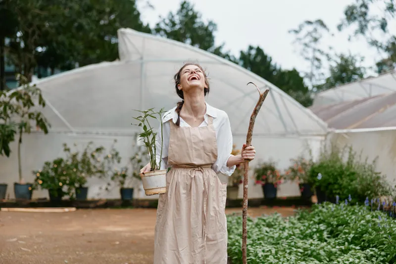 Joyful woman in a garden holding a potted plant and gardening tools, showcasing a connection with nature and sustainable living