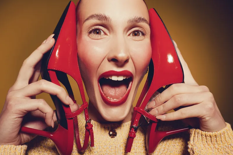 Happy woman with red lipstick holds red high heel shoes near face, smiling with open mouth. Close-up portrait with warm lighting and beige sweater. Concept of fashion and joy.