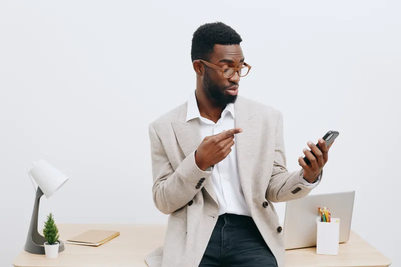 Professional young man with glasses in formal attire using smartphone while standing in modern office environment, white background, workspace atmosphere, technology, communication, business concept