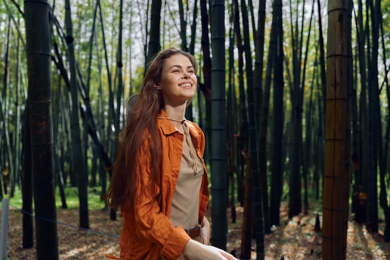Woman in bamboo forest with longhair and smile, wearing orange jacket and walking through tall stalks, enjoying nature and sunlight for a serene outdoor portrait.