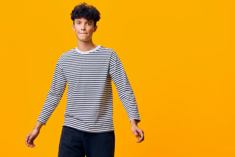 young man in striped shirt standing relaxed against bright orange background, casual style with thoughtful expression and natural curly hair