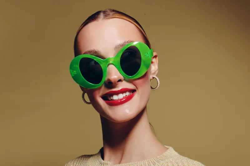 Smiling woman wearing large green sunglasses and red lipstick poses against beige background. Fashion portrait with bright makeup and trendy accessories. Concept of style and confidence.