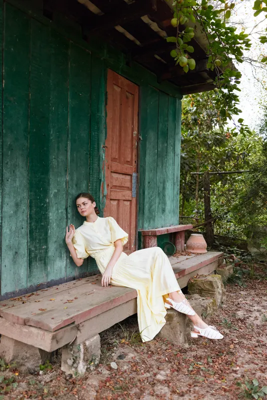 Woman in a pale yellow dress relaxes on a wooden bench outside a rustic green house, posing gracefully as afternoon light frames a tranquil countryside scene.
