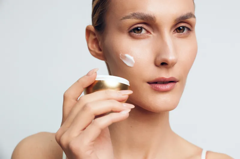 Young woman applying white face cream on cheek holding jar with golden lid. Close-up portrait with natural makeup on light background. Concept of skincare and beauty routine.