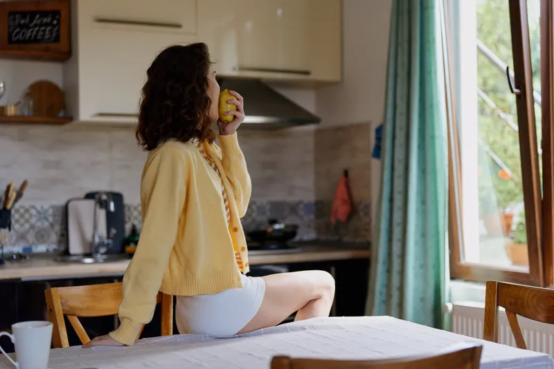 A woman in a yellow cardigan sits at a kitchen island, sipping from a cup as morning light fills a cozy home with casual, relaxed atmosphere, quiet kitchen sounds, simple routine, gentle morning