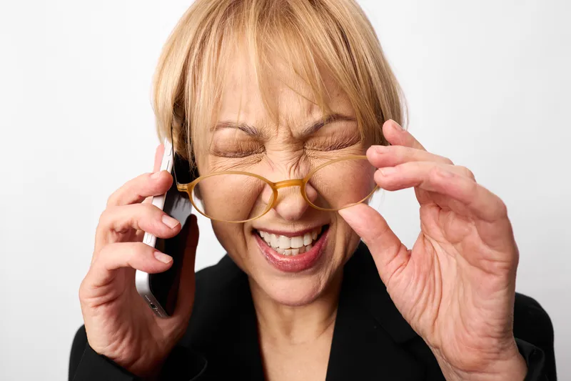 Senior woman in business suit laughing while using smartphone, lifting sunglasses with fingers, playful expression, white studio background, close up portrait, eyewear fashion