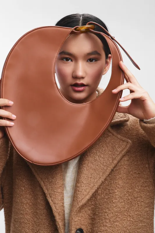 asian woman poses with a bold brown sculptural accessory in a studio, showcasing contemporary fashion and confident poise, creating a striking modern portrait.