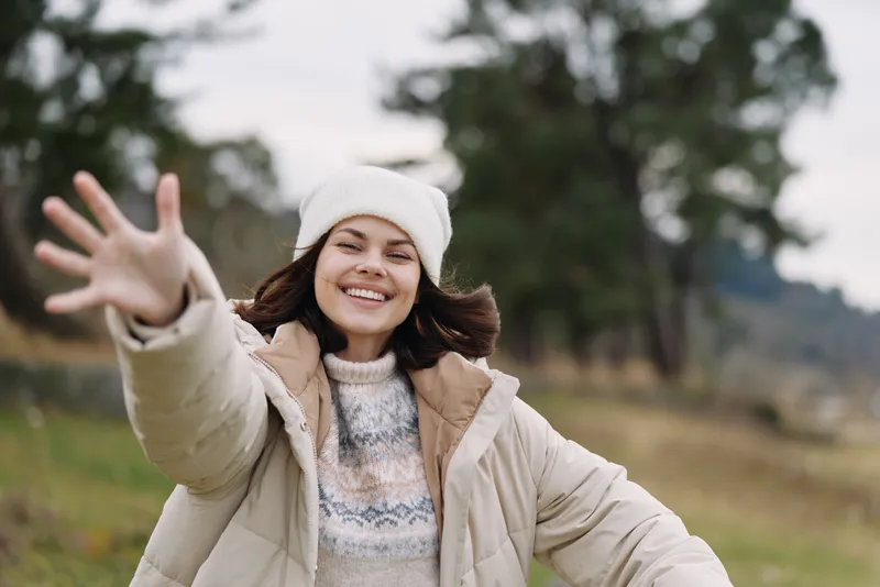 Woman in a warm coat and hat outdoors, smiling with open arms in a sunny natural setting, winter scene, carefree mood and fresh air.