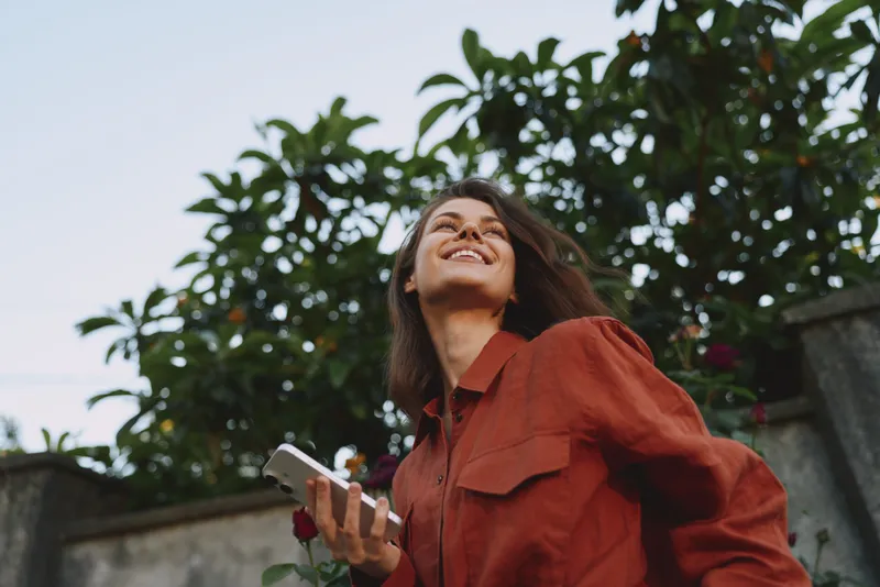 Smiling woman with long hair in a red shirt outdoors, looking up, holding a smartphone. Bright natural daylight, lush greenery, casual lifestyle moment, carefree mood, candid portrait