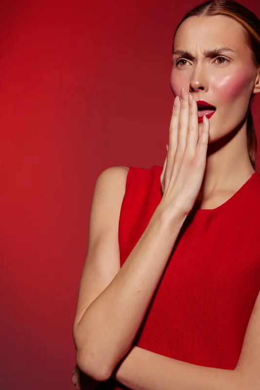 Surprised woman in red dress covers mouth with hand, looking away on red background. Concept of shock, emotion, and fashion portrait with dramatic lighting and makeup.